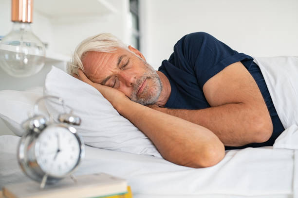 An older man sleeping in bed with an alarm clock on the bedside table.