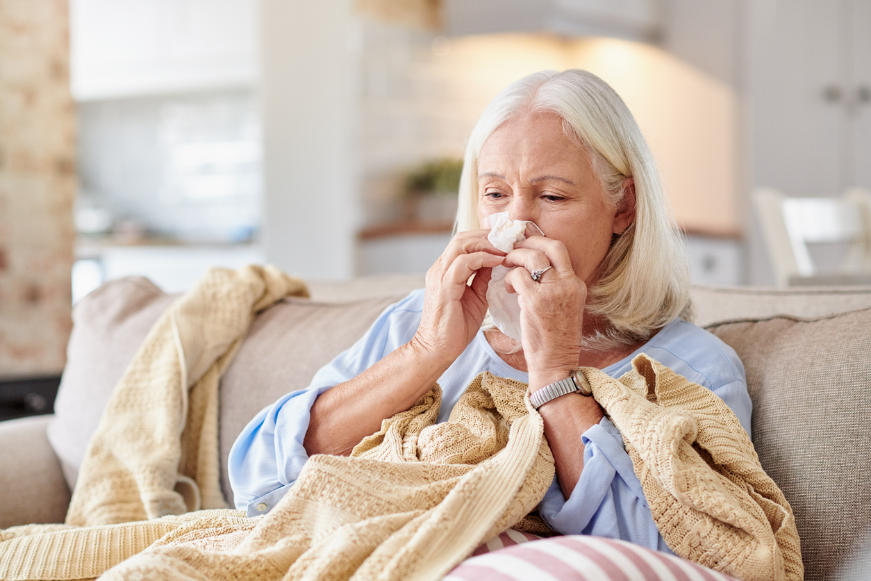 Elderly woman blowing her nose into a tissue while wrapped in a blanket