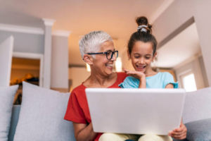 A senior woman and her young granddaughter smile while using a tablet together.